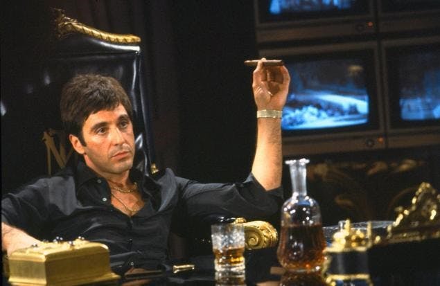 Al Pacino i huvudrollen i Scarface sittande bakom ett skrivbord med en cigarr i handen.